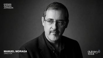 Black-and-white studio portrait of Manuel Moraga, director, wearing glasses and a dark blazer, looking slightly to the side against a dark plain background, with “Godwin Austen Johnson” in the top left and “dubaieye 103.8” in the bottom right.