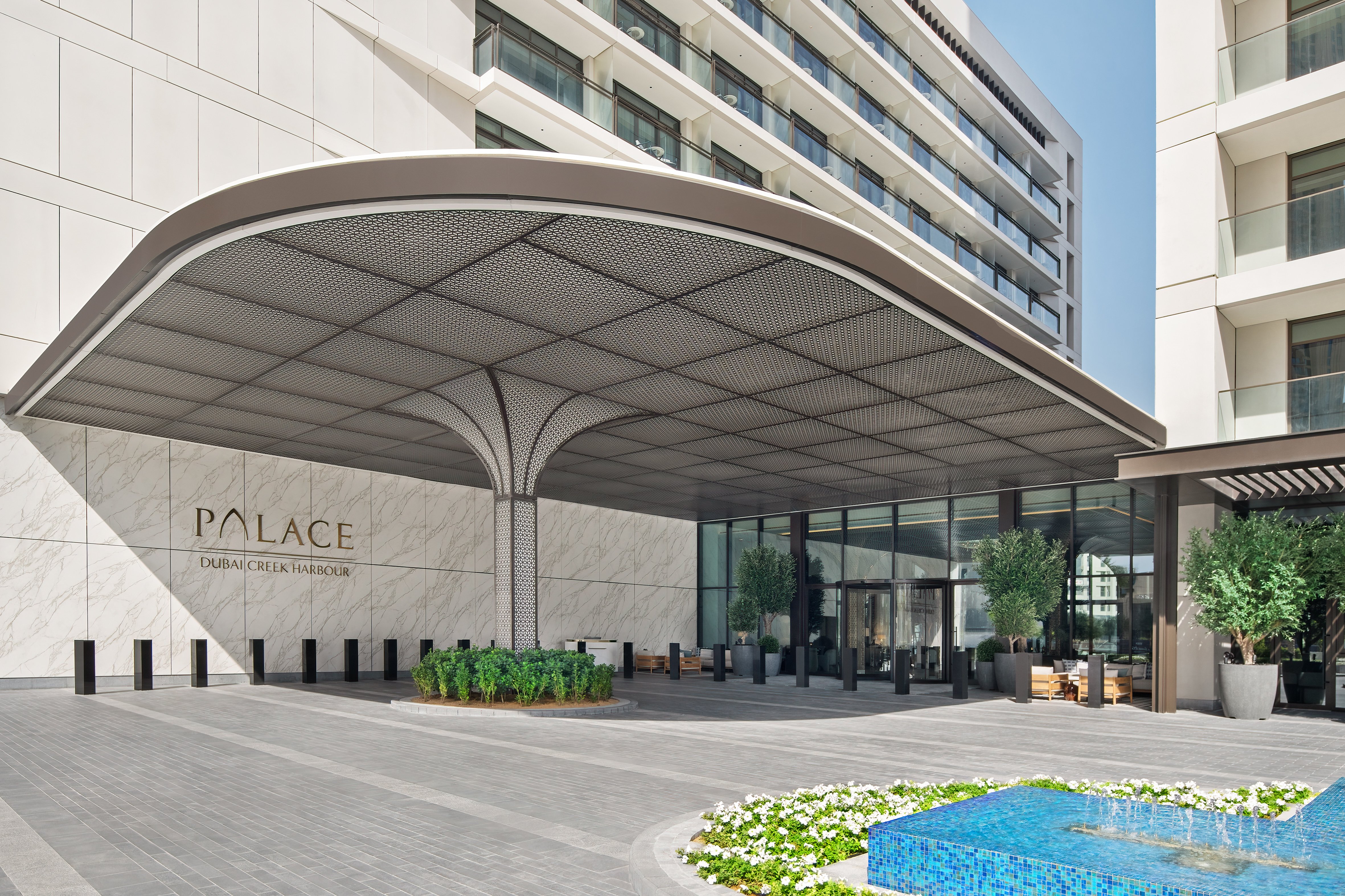 Elegant main entrance of Palace Dubai Creek Harbour hotel, featuring a modern curved canopy, marble facade, and landscaped surroundings with a decorative water feature.